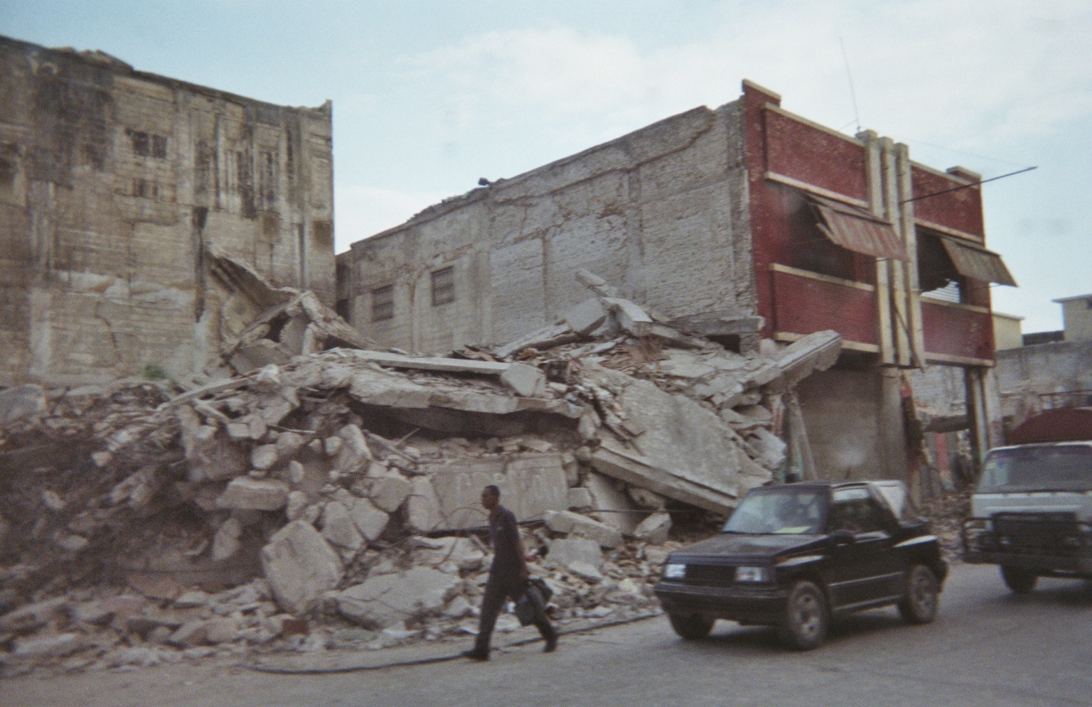 <em><strong>Commuters pass by a collapsed building, a common sight in Port-au-Prince, Haiti (Photo courtesy Dr. Hélène Rousseau)</em></strong>